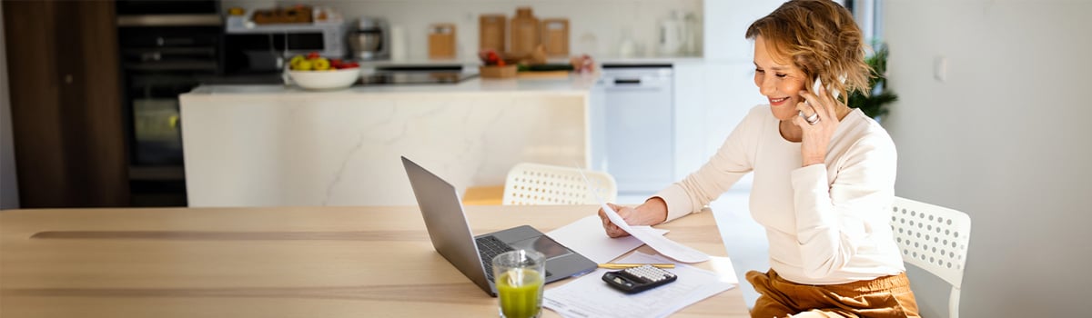 woman on the phone, looking at document with laptop open in her kitchen